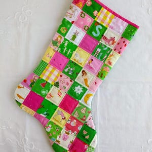May include: A quilted Christmas stocking featuring a patchwork design of various fabric squares in shades of bright pink, lime green, and white. The fabric squares display diverse patterns including holiday motifs, animals, and a prominent letter S.