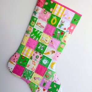 May include: A patchwork Christmas stocking constructed from various fabric squares in shades of pink, green, yellow, and white. The squares feature diverse printed patterns including animals, flowers, holiday motifs, and a yellow letter O.