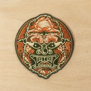May include: A round embroidered patch with a green outline and a brown, furry creature with a fierce expression. The creature has a large, open mouth with sharp teeth and a prominent nose.