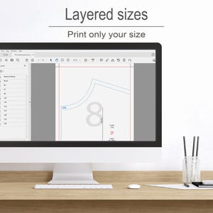 a computer monitor with a graphic design program open, showing a layered design with various sizes and shapes. The monitor is placed on a wooden desk, and there are some pens and a cup nearby.