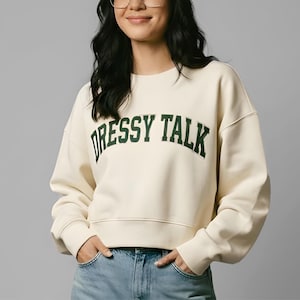 May include: Cream-colored sweatshirt with the text "DRESSY TALK" in dark green block letters. The sweatshirt has a crew neck and long sleeves. The model is wearing blue jeans.