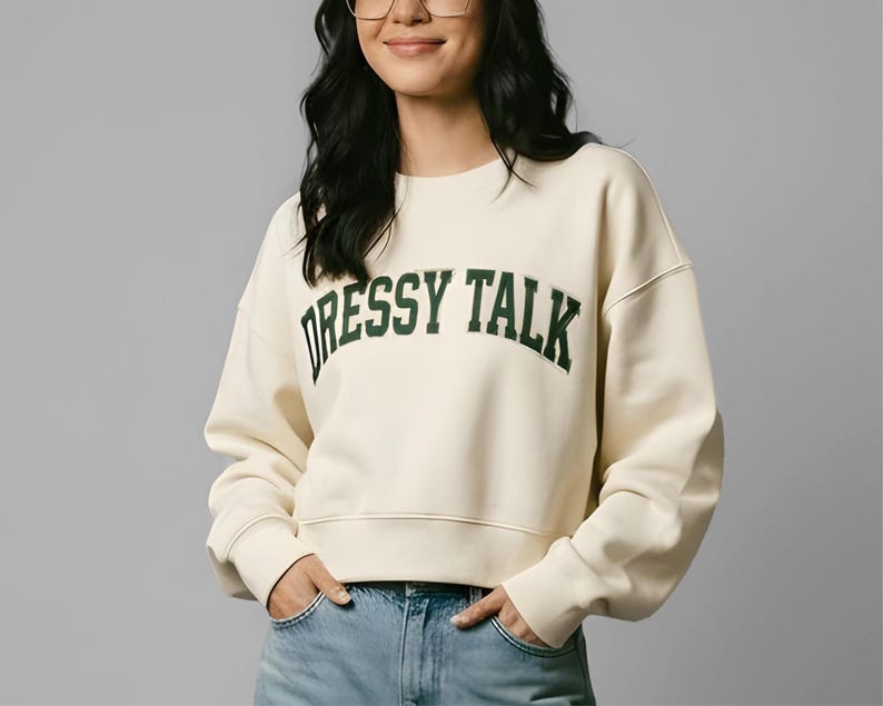 May include: Cream-colored sweatshirt with the text "DRESSY TALK" in dark green block letters. The sweatshirt has a crew neck and long sleeves. The model is wearing blue jeans.