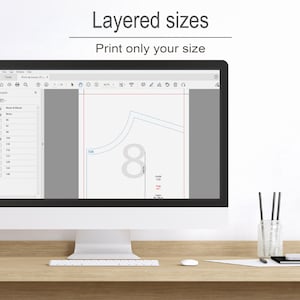 a computer monitor with a graphic design program open, showing a layered design with various sizes and shapes. The monitor is placed on a wooden desk, and there are some pens and a cup nearby.