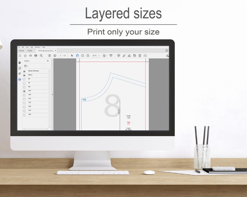 a computer monitor with a graphic design program open, showing a layered design with various sizes and shapes. The monitor is placed on a wooden desk, and there are some pens and a cup nearby.