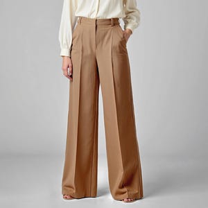 May include: Tan wide-leg trousers with a high waist and a tailored fit. The pants have a classic design with a clean, minimalist aesthetic. The trousers are paired with a cream-colored long-sleeve shirt.