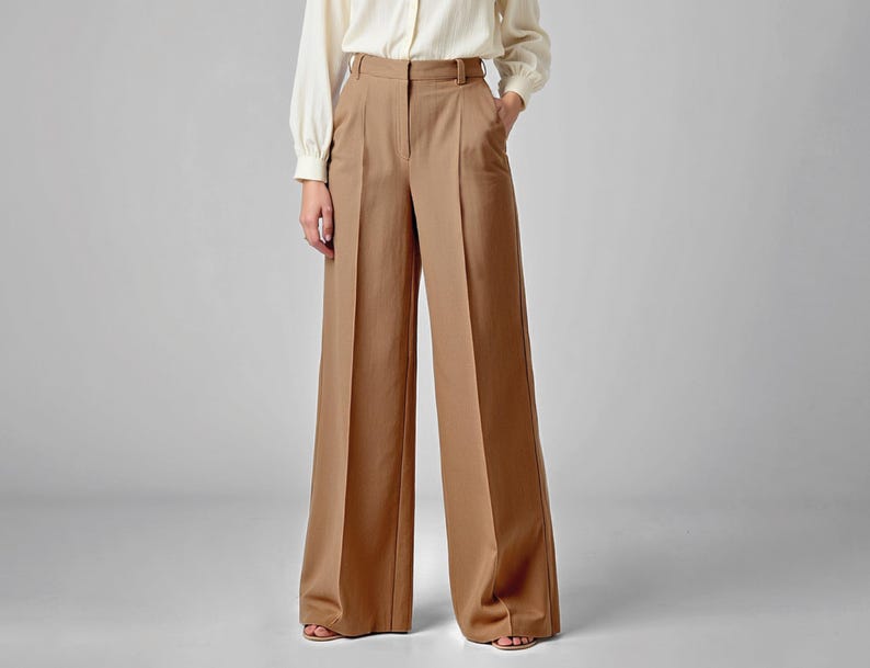 May include: Tan wide-leg trousers with a high waist and a tailored fit. The pants have a classic design with a clean, minimalist aesthetic. The trousers are paired with a cream-colored long-sleeve shirt.