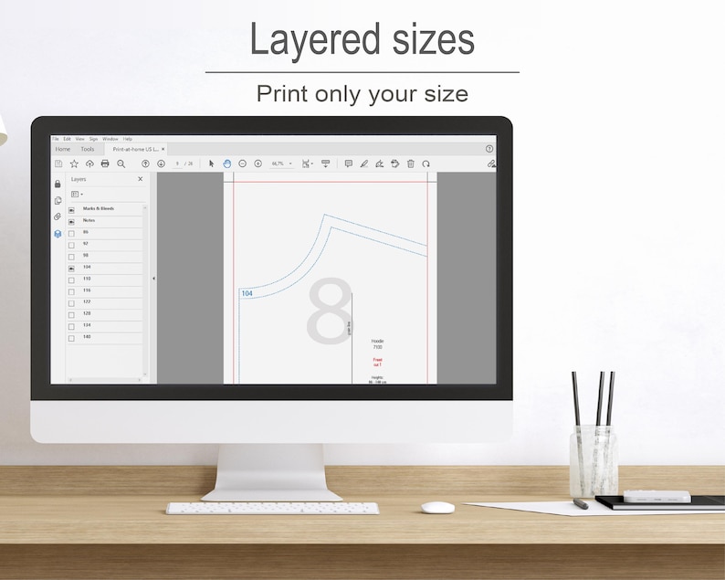 a computer monitor with a graphic design program open, showing a layered design with various sizes and shapes. The monitor is placed on a wooden desk, and there are some pens and a cup nearby.