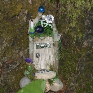 Puede incluir: Una casa de hadas en miniatura hecha de ramitas y corteza, con una pequeña puerta, un techo de musgo verde y decoraciones azules y blancas.