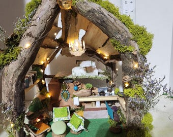 May include: A miniature fairy house made of wood branches and moss, with a green grass floor, a small couch, and a chandelier hanging from the ceiling.