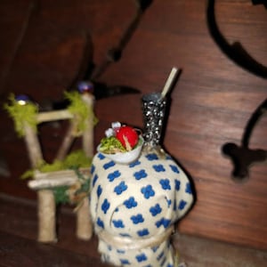 May include: A miniature dollhouse scene with a small chair and a table set with a plate of food and a drink. The table is covered in a blue and white patterned cloth.
