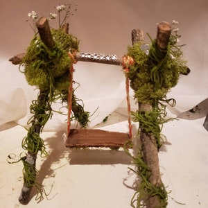 May include: A miniature wooden swing set with a brown wooden seat, decorated with moss and small white flowers. The swing is suspended from two branches with red and white string.