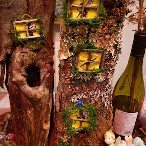 May include: Two miniature fairy houses made from tree trunks with windows and moss accents. The houses are decorated with fairy wings and are sitting on a bed of moss.