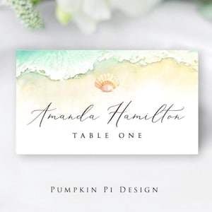 May include: A rectangular place card with a watercolor beach scene. The top of the card features a turquoise ocean meeting a sandy shore, with a seashell. The name "Amanda Hamilton" is written in elegant script, with "TABLE ONE" below. "PUMPKIN PI DESIGN" is at the bottom.