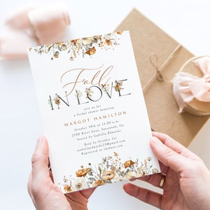 Pode incluir: Um convite para chá de panela com o texto "Fall in Love" em escrita elegante. O convite apresenta detalhes florais em cores quentes de outono e inclui detalhes do evento, como data, hora e local.