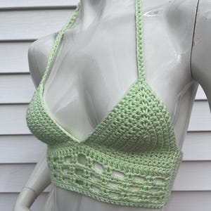 May include: A light green, handmade crochet halter top. The top features a triangle-shaped bust with a halter neck tie and a decorative openwork design along the bottom edge. The garment is displayed on a white mannequin.