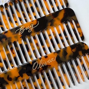 May include: Close-up of several personalized combs. The combs have a tortoiseshell pattern in shades of brown, black, and amber. Each comb is inscribed with a name in gold script: Sabrina, Bug, and Olivia. The combs have evenly spaced teeth.