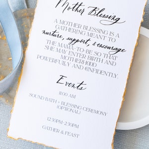 May include: A white card with gold edges and black text. The card reads "Mother Blessing" and describes a gathering meant to nurture, support, and encourage the mother-to-be. The card lists events for the gathering, including a sound bath and blessing ceremony, and a gather and feast.
