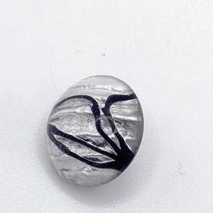 May include: A clear, oval-shaped glass bead with a silver interior and black, abstract linear design. The bead is smooth and reflective, with the black lines creating a contrasting pattern against the silver background.