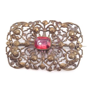 May include: A vintage gold-toned brooch with a delicate filigree design and a large, faceted, red gemstone in the centre.