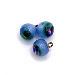 May include: Three round, light blue glass beads with colourful swirls of green, blue, and red. Each bead has a small, gold-coloured loop attached. The beads are scattered on a white background.