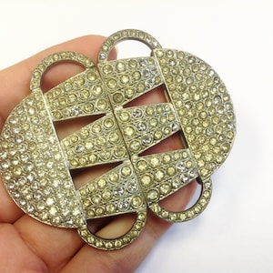 May include: A silver-toned metal belt buckle with a geometric design. The buckle is covered in small, clear rhinestones.