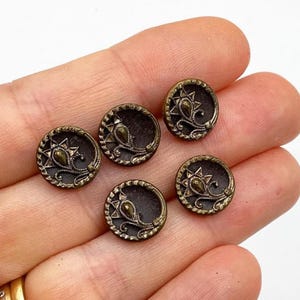 May include: Five antique brass buttons with a dark brown center and ornate gold-colored designs. Each button features a starburst and a stylized teardrop shape, perfect for sewing or crafting projects.