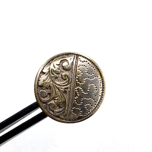 May include: A vintage, circular button with an elaborate design. One half displays a detailed floral pattern, the other a textured, dotted design. The button is held by tweezers against a white backdrop, highlighting its metallic colour and intricate details.