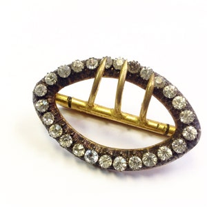May include: An oval buckle in gold tone with a decorative design of small clear rhinestones around the edge. The buckle has a metal bar with three prongs.