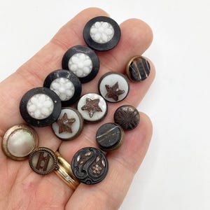 May include: A collection of vintage buttons in various colors and designs, including black, white, brown, and gold. The buttons feature different patterns, such as flowers, stars, and swirls.