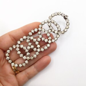 May include: A vintage silver-toned brooch with a series of interconnected, open circles. Each circle is encrusted with clear rhinestones. The brooch is held against a white background, showcasing its intricate design.