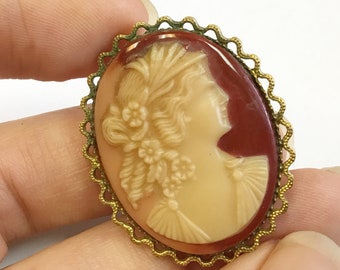 May include: A gold-tone cameo brooch with a white cameo of a woman's profile with a floral design. The cameo is set in a red oval background.