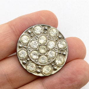 May include: A round, silver-toned button with a central cluster of clear rhinestones surrounded by two concentric circles of smaller rhinestones. The button has a vintage aesthetic and is likely for sewing or embellishment.