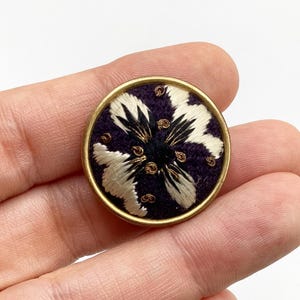 以下が含まれることがあります： 紫色のベルベットの中心と白と黒の刺繍の花のデザインが施された、丸い金色のボタンです。
