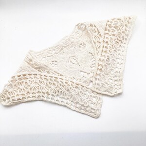 May include: Cream-coloured, open-weave crochet shawl. The triangular shawl features intricate floral and geometric patterns. The delicate, lightweight design is visible against the white background. The shawl is folded, showcasing the detailed craftsmanship.