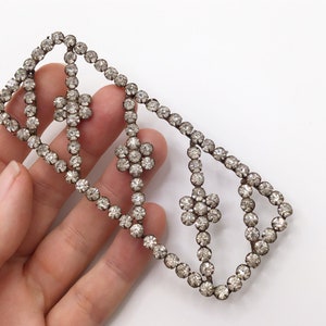 May include: A silver rhinestone trim with a geometric design. The trim features a series of connected loops and flower-shaped clusters of rhinestones.