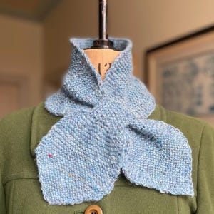 Könnte beinhalten: Hellblauer Strickschal mit bunten Flecken, auf einer Schaufensterpuppe präsentiert. Der Schal hat eine einzigartige Form, mit einem Kragen und zwei ausgestellten Enden. Der Schal ist aus einem weichen, strukturierten Garn gefertigt, ideal für Wärme und Stil.