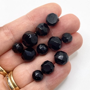 May include: A collection of black faceted glass beads, some round and some with a more geometric shape. The beads are arranged on a hand.