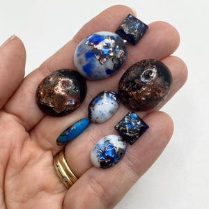 May include: A selection of handmade glass beads in diverse forms. The beads display a blend of colours, such as blue, white, and copper, with a speckled or marbled appearance. Some are oval, others are square or cylindrical in shape.