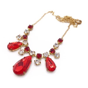 May include: A gold-coloured necklace with red and clear crystal accents. The necklace features a delicate chain and a series of red teardrop-shaped crystals, interspersed with smaller red and clear crystals.