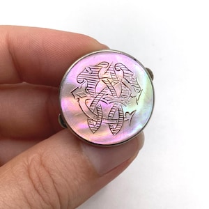 May include: A round, iridescent mother-of-pearl button with a silver-tone metal frame. The button features an engraved, stylized design in a swirling pattern. The button is held by a hand against a white background.