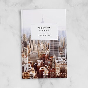 May include: A white book with a cityscape photo on the cover. The title "Thoughts & Plans" is printed in black text on a white rectangle. The author's name, "Tommy Smith", is printed below the title.