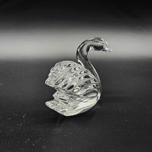 May include: A clear glass swan figurine with detailed feather and wing patterns. The swan is posed with its head held high, and its body is curved. The figurine is displayed on a dark surface.