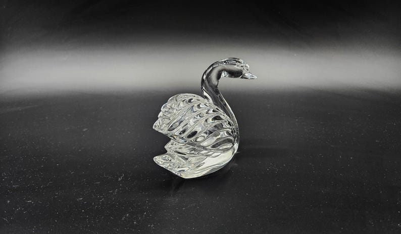 May include: A clear glass swan figurine with detailed feather and wing patterns. The swan is posed with its head held high, and its body is curved. The figurine is displayed on a dark surface.