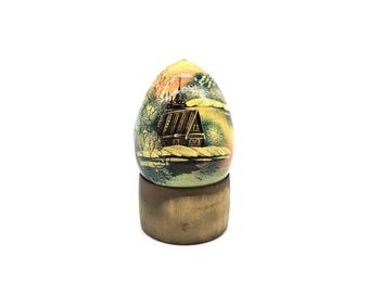 May include: A decorative egg-shaped ornament with a painted landscape scene featuring a wooden building. The egg is set on a wooden stand. The color palette includes shades of yellow, orange, green, and brown.