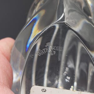 May include: Clear crystal decorative object with a unique, curved design. The object has a smooth, reflective surface that catches the light, creating a rainbow effect. The word "Waterford" is etched into the crystal. The object is held by a hand.