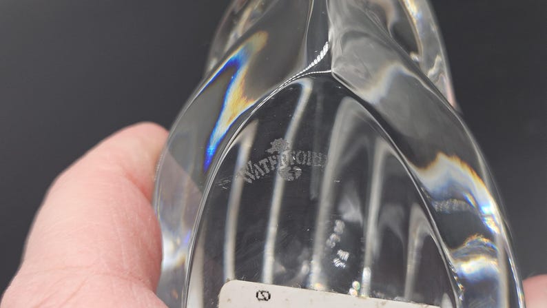 May include: Clear crystal decorative object with a unique, curved design. The object has a smooth, reflective surface that catches the light, creating a rainbow effect. The word "Waterford" is etched into the crystal. The object is held by a hand.