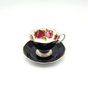 Può includere: Un set di tazza e piattino da tè neri con finiture dorate. L'interno della tazza presenta un motivo floreale con rose rosse e rosa. La tazza e il piattino sono su sfondo bianco.