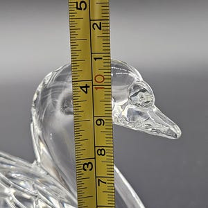 May include: A clear glass duck figurine with a textured surface. The duck's head is in focus, with a yellow ruler showing a height of approximately 4 inches. The figurine is on a gray background.