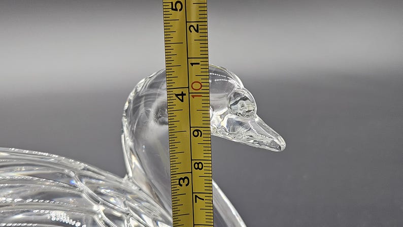 May include: A clear glass duck figurine with a textured surface. The duck's head is in focus, with a yellow ruler showing a height of approximately 4 inches. The figurine is on a gray background.