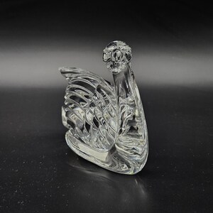 May include: A clear glass swan figurine with intricate detailing. The swan is positioned on a dark surface, showcasing its elegant form. The design features a detailed head and a textured wing, creating a decorative piece.